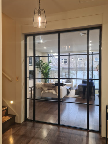 Modern black metal double door with side panels opening to a bright living room with beige sofas and wooden floor