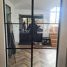 Double hinged black metal frame glass doors opening to a modern kitchen with chevron wooden floor