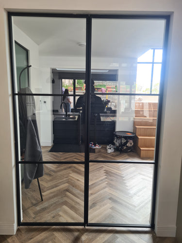 Double hinged black metal frame glass doors opening to a modern kitchen with chevron wooden floor