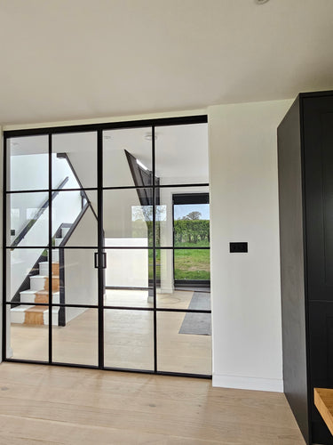 double hinged black metal framed glass doors partitioning a bright room with wooden stairs and light wood flooring