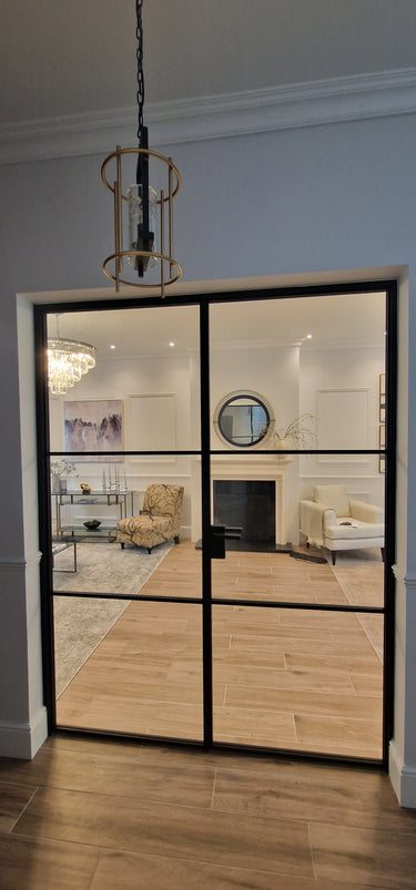 Double hinged glass door with black metal frame opening to modern living room with chandelier, armchairs, and fireplace