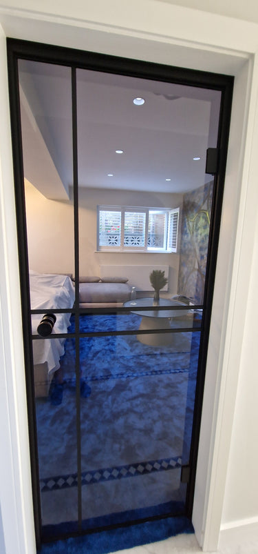 Modern hinged glass door with black frame opening to blue carpeted living room with sofa and round table