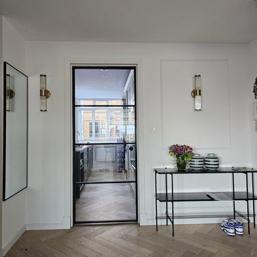 Modern interior with black framed hinged glass door leading to kitchen, white walls, console table with vases and flowers