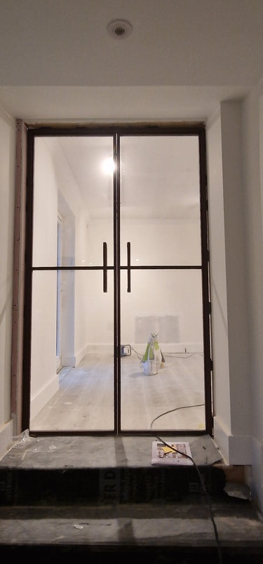 Double-hinged glass door with black metal frame in a white room under renovation