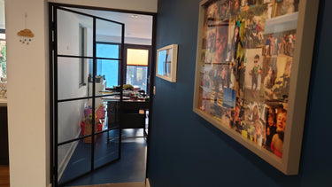 Interior view of a modern home office with a black-framed glass hinged door and a photo collage on a blue wall