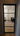 Black framed hinged glass door opening to room with wooden floor and black armchair with patterned cushion