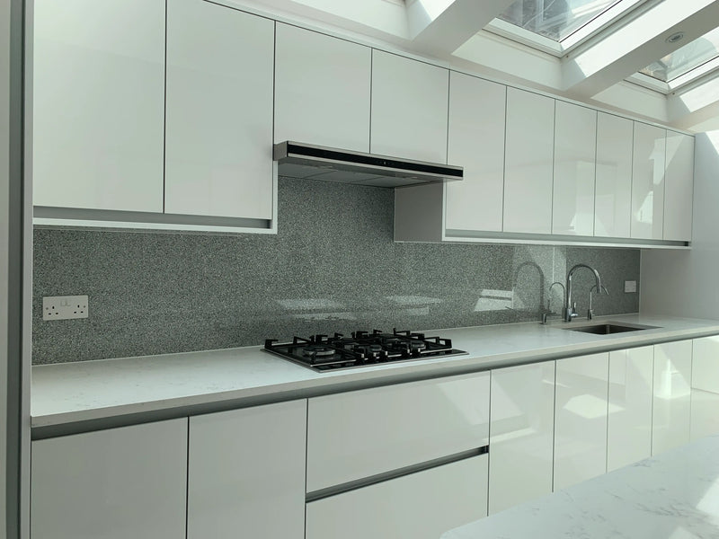 Modern kitchen with white cabinets, marble countertops, gas stove, gray backsplash, and dual sink under skylight