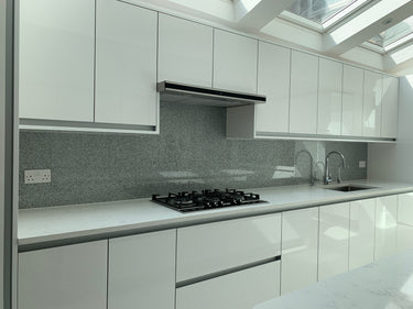 Modern kitchen with white cabinets, marble countertops, gas stove, gray backsplash, and dual sink under skylight