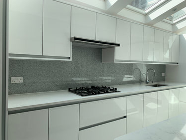 Modern kitchen with white cabinets, marble countertops, gas stove, gray backsplash, and dual sink under skylight