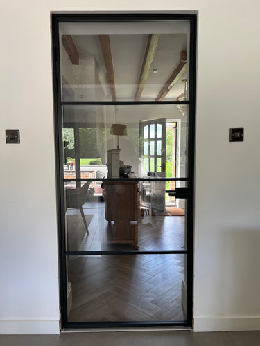 Black framed glass interior door showing a modern home with wooden furniture and outdoor garden view