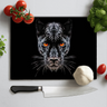 Black chopping board with orange-eyed panther face design, kitchen knife, garlic, tomatoes, and basil on white surface