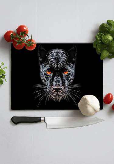Black chopping board with orange-eyed panther face design, kitchen knife, garlic, tomatoes, and basil on white surface