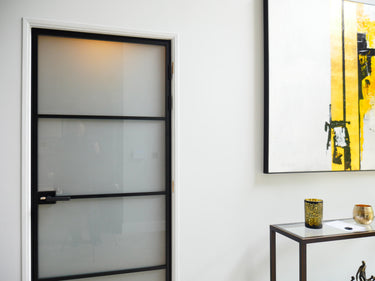 Modern interior with frosted glass hinged door framed in black metal, white walls, and abstract yellow art