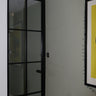 Black framed hinged glass door in modern room with gray wall, radiator, and yellow framed artwork