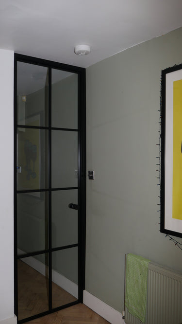 Black framed hinged glass door in modern room with gray wall, radiator, and yellow framed artwork