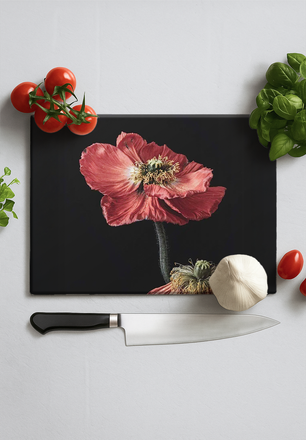 Black chopping board with red poppy flower design, surrounded by cherry tomatoes, garlic, basil, and a chef's knife