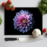 Black chopping board with large purple dahlia print surrounded by tomatoes, garlic, basil, and chef knife on white surface