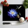 Black chopping board with blue lotus flower and dragonfly design surrounded by tomatoes, garlic, basil, herb, and knife