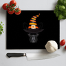 Black chopping board with a black pug wearing an orange and black striped witch hat, surrounded by kitchen items