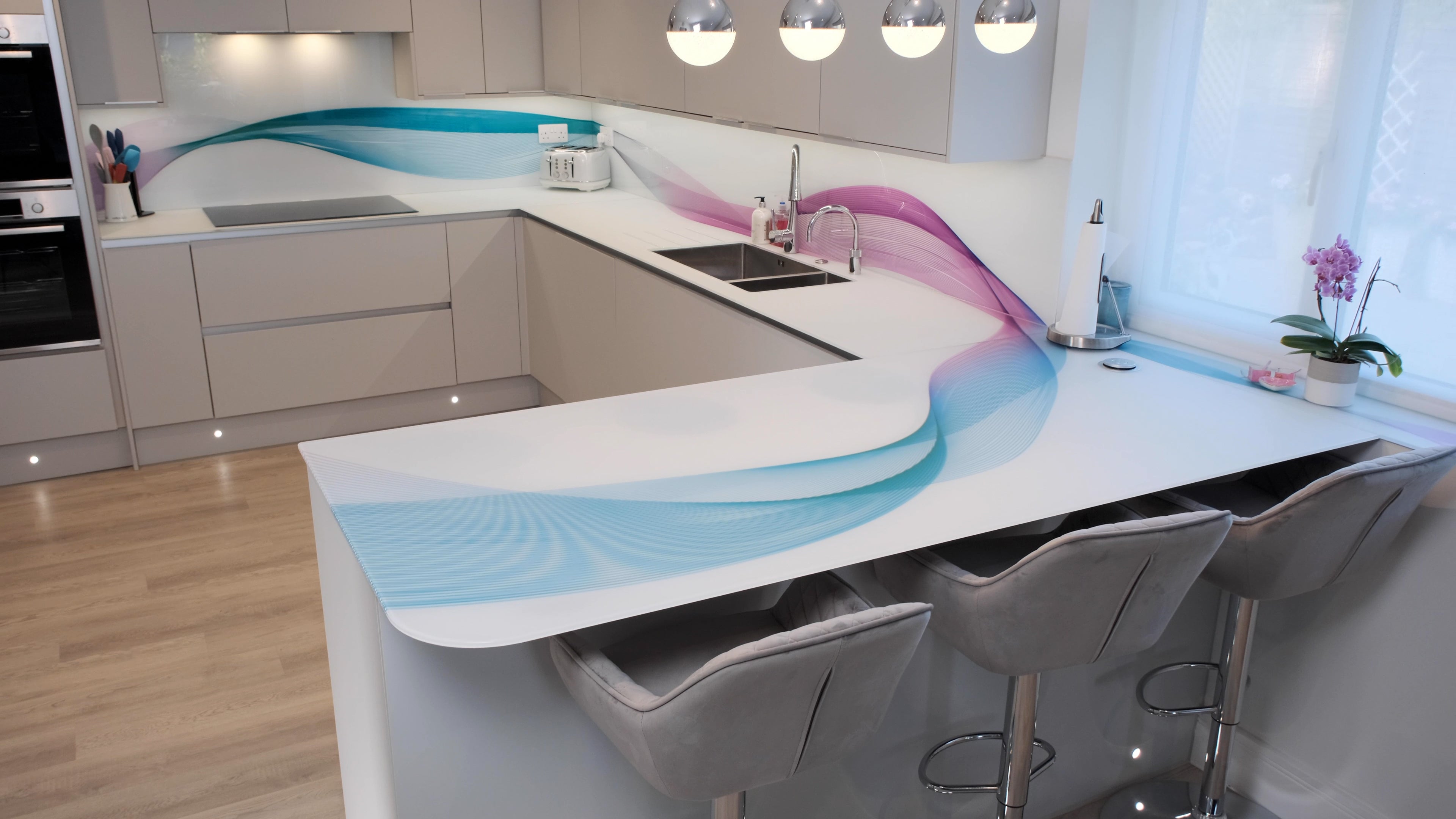 Modern kitchen with bespoke glass splashback featuring blue and pink wave design, sleek white countertops, and bar stools.