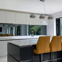 Modern kitchen with sleek white cabinets, black island, and glass splashback, featuring orange bar stools and pendant lights.