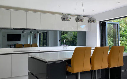 Modern kitchen with sleek white cabinets, black island, and glass splashback, featuring orange bar stools and pendant lights.