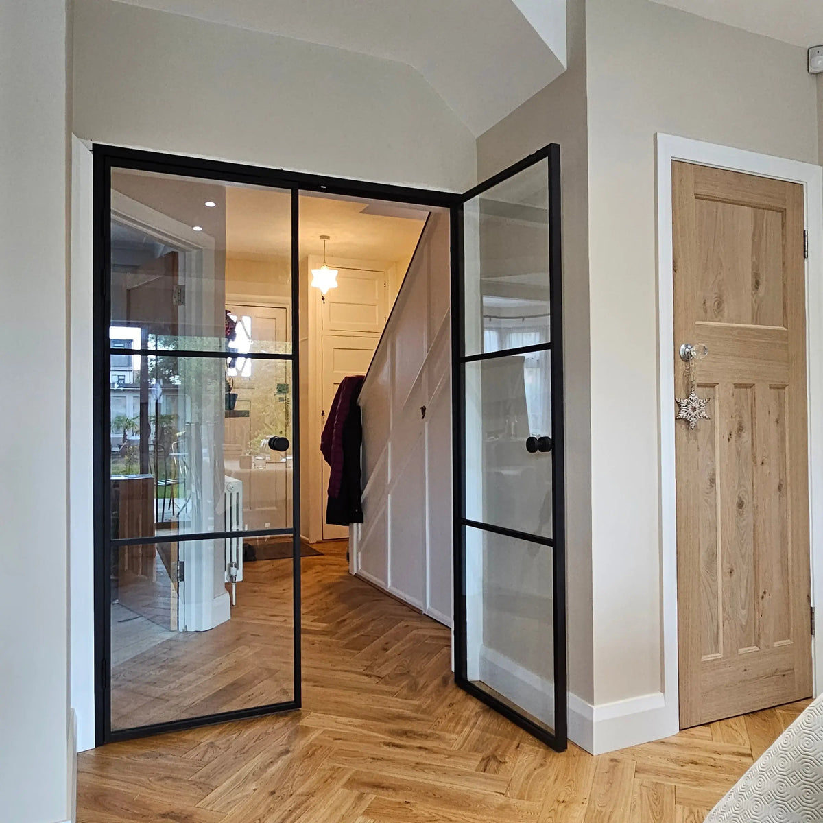 Black-framed glass doors with grid design, opening to hallway with herringbone wood floor and staircase