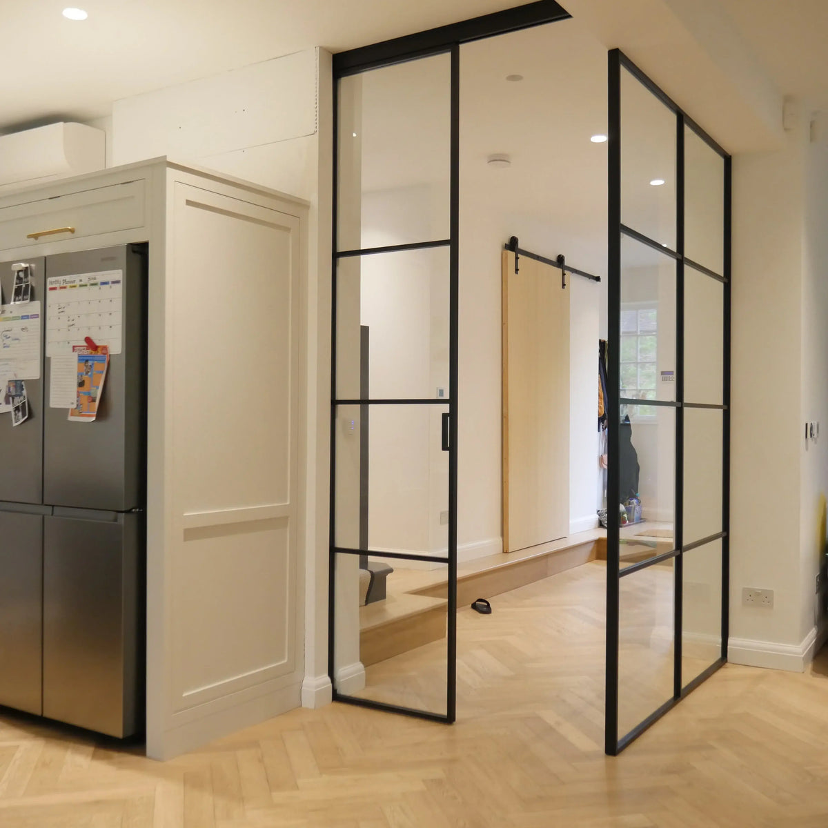 Black-framed glass partition with sliding door in modern kitchen, featuring herringbone wood flooring and sleek cabinetry.
