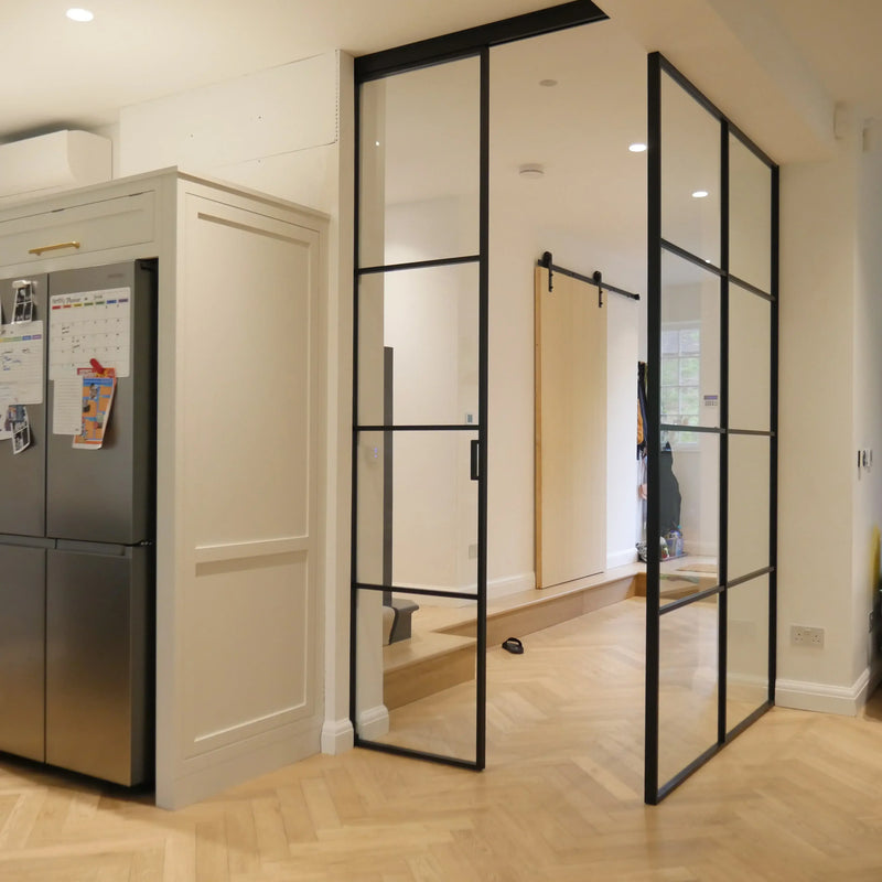 Black-framed glass partition with sliding door in modern kitchen, featuring herringbone wood flooring and sleek cabinetry.