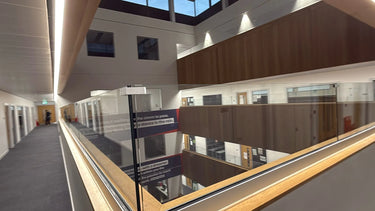 An interior architectural view of a modern multi-story building atrium with glass railings, wood paneling, and a hallway on the left.