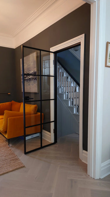 Black framed glass hinged door opening to staircase, with mustard velvet sofa in gray living room