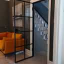 Modern hinged black metal frame glass door open to staircase, with dark walls and mustard yellow sofa