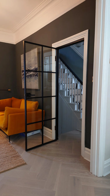Modern hinged black metal frame glass door open to staircase, with dark walls and mustard yellow sofa