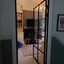 Hinged black-framed glass door opens to a cozy living room with chandelier, fireplace, and bookshelves