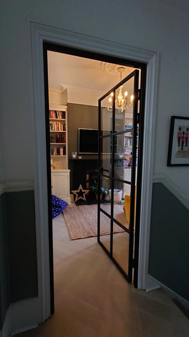 Hinged black-framed glass door opens to a cozy living room with chandelier, fireplace, and bookshelves