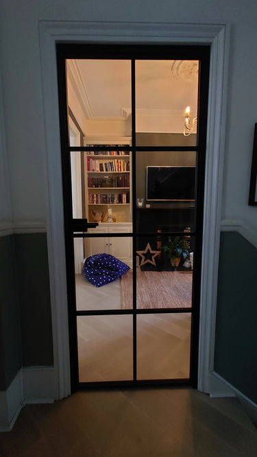 Black framed glass hinged interior door revealing a cozy living room with bookshelf, TV, and blue star-patterned bean bag