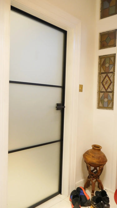Frosted glass hinged door with black frame in a white wall hallway, stained glass panels, shoes on floor