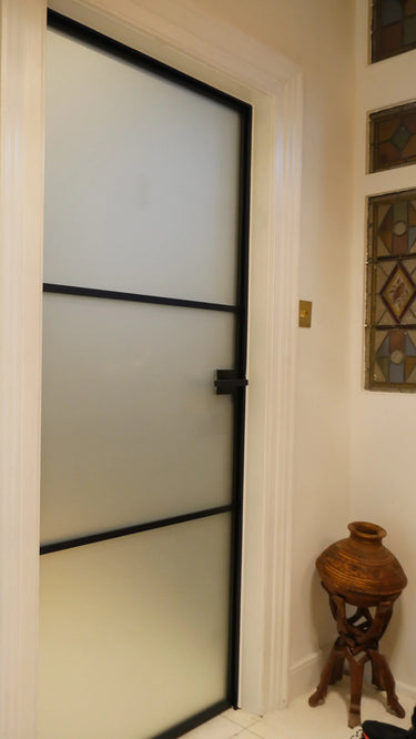 Modern frosted glass hinged door with black frame in a white interior hallway featuring stained glass and wooden decorative vase stand